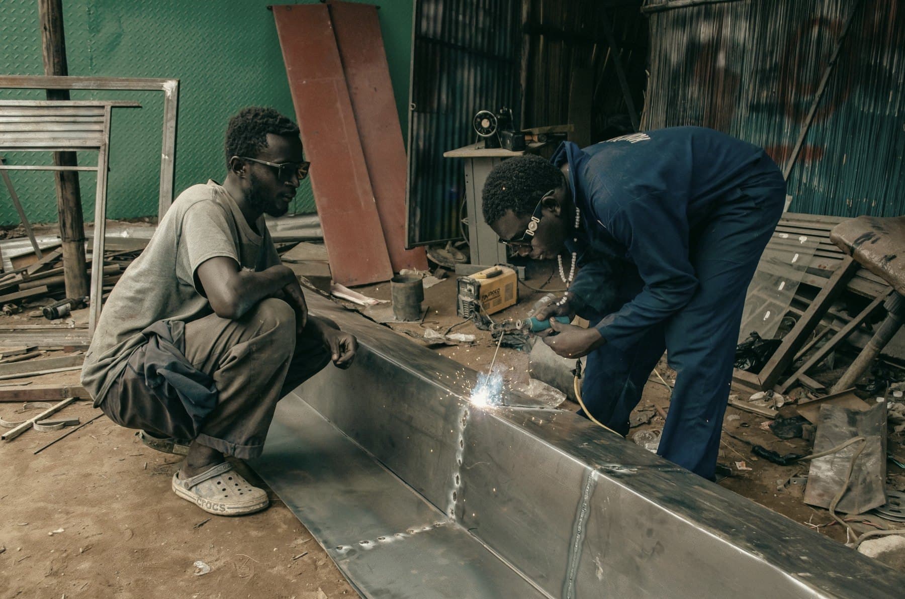 Two men in a metal workshop, one inspecting the work while the other welds